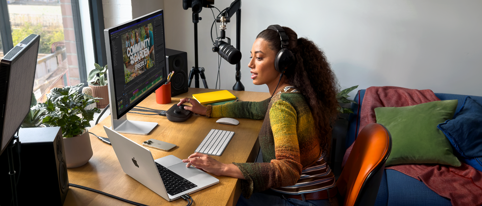 Eine Frau arbeitet bei sich zu Hause an ihrem Schreibtisch mit einem MacBook Pro, das an zwei externe Displays angeschlossen ist. Sie trägt Kopfhörer und spricht in ein Mikrofon